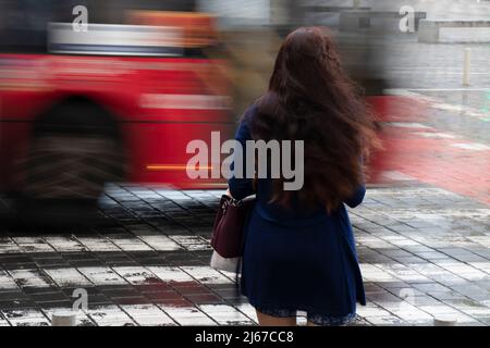 Jeune femme aux longs cheveux ondulés attendant sur un carrefour par temps pluvieux avec circulation en mouvement flou, vue arrière Banque D'Images