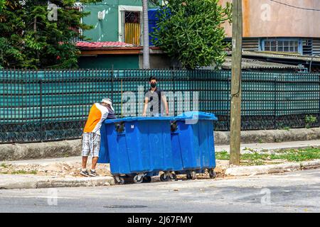 Un homme cubain cherche quelque chose dans les poubelles de la ville. Un autre homme avec masque facial protecteur marche dans le trottoir observant la même scène. Banque D'Images