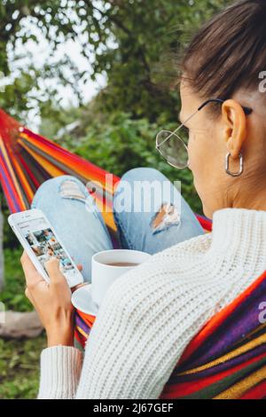 Joli créateur de contenu dans hamac utiliser smartphone regarder l'écran de chat dans les médias sociaux boire thé lire les nouvelles en ligne navigation photo en vacances, vue arrière Banque D'Images