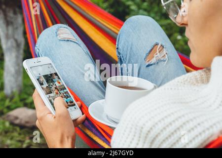 Joli créateur de contenu dans hamac utiliser smartphone regarder l'écran de chat dans les médias sociaux boire thé lire les nouvelles en ligne navigation photo en vacances, vue arrière Banque D'Images