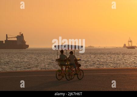 Deux garçons dans un vélo à quatre roues (vélo de surrey) à la promenade regardant un navire dans la mer au coucher du soleil. Banque D'Images