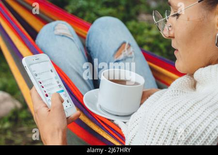 Joli créateur de contenu dans hamac utiliser smartphone regarder l'écran de chat dans les médias sociaux boire thé lire les nouvelles en ligne navigation photo en vacances, vue arrière Banque D'Images