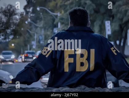 L'agent FBI mâle porte un manteau bleu foncé avec le logo FBI regardant dans la rue avec des voitures au crépuscule vu de derrière. Banque D'Images