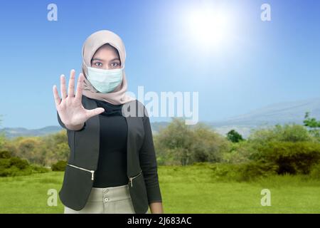 Femme musulmane asiatique portant un foulard et un masque de visage avec un signe d'arrêt sur fond bleu ciel Banque D'Images