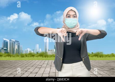 Femme musulmane asiatique portant un foulard et un masque de visage debout montrant le signe du pouce vers le bas sur fond bleu ciel Banque D'Images