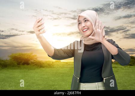 Femme musulmane asiatique dans un foulard emportant un selfie avec son téléphone portable avec un fond de coucher de soleil Banque D'Images