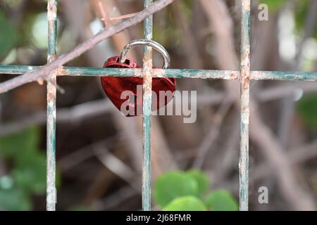 Cadenas rouge en forme de coeur brillant, verrouillé autour d'une clôture en fil de fer, avec vue de l'orifice de la clavette depuis le dessous de la serrure Banque D'Images