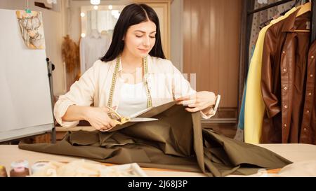 couturière aux cheveux noirs en atelier, portrait au travail. Table avec tissu et outils de couture, à l'intérieur. Coupes avec ciseaux Banque D'Images