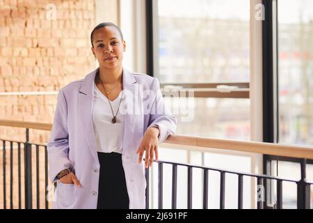 Portrait d'une femme adulte de taille moyenne LGBTQ multiraciale confiante, penchée sur une rampe dans un espace de bureau moderne Banque D'Images