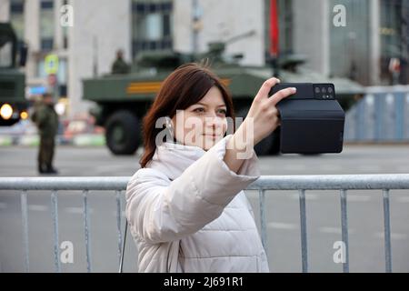 Bonne fille prenant des photos de selfie sur smartphone sur fond de véhicules blindés des forces militaires russes lors d'une répétition de la parade du jour de la victoire Banque D'Images