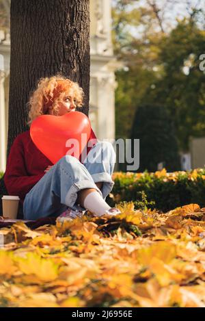 Être amoureux concept. Photo verticale en plein air d'une adolescente moderne aux cheveux bouclés tenant un ballon en forme de cœur tout en étant assise sous un arbre sur des feuilles d'automne jaunes dorées. Photo de haute qualité Banque D'Images