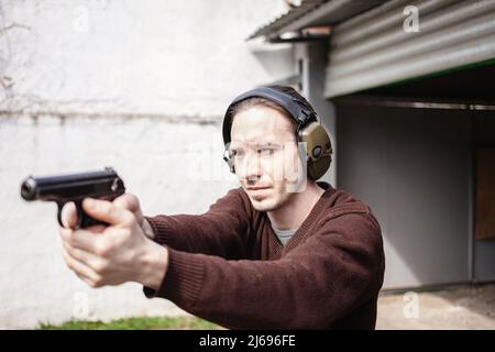 Un jeune homme cherche un fusil. Un homme portant un casque de protection. Gamme de tir de pneu extérieur. Hobby. Arme à feu contre le mur blanc Banque D'Images