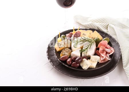 Carte de charcuterie avec assortiment de fromages, salami et prosciutto servis avec des olives et un verre de vin rouge sur une assiette noire sur fond de table blanc. Banque D'Images