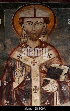 Saint évêque berce une Bible dans son bras gauche et fait un geste de bénédiction avec sa main droite. Fresque médiévale, peinte dans le style lombardique. Au début des années 1300 Chiesa di San Giorgio (église Saint-Georges) à Varenna, près du lac de Côme en Lombardie, Italie. Banque D'Images