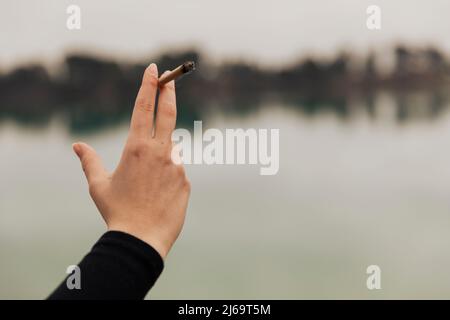 Main d'une jeune fille tenant une cigarette de marijuana dans un lac à l'extérieur Banque D'Images