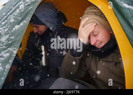 Les gars sont assis dans une tente, préparant la nourriture pendant une randonnée d'hiver. Près de la tente dans la neige se trouve leur équipement. Deux hommes mangent dans la tente. C Banque D'Images