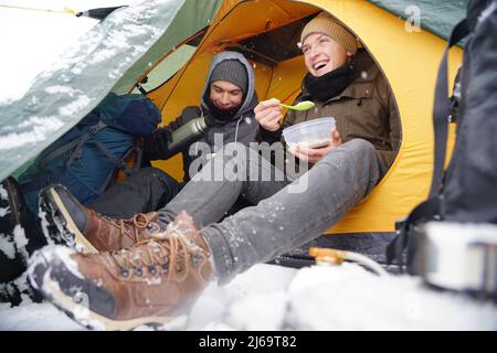 Les gars sont assis dans une tente, préparant la nourriture pendant une randonnée d'hiver. Près de la tente dans la neige se trouve leur équipement. Deux hommes mangent dans la tente. C Banque D'Images