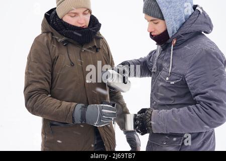 il boit le thé chaud des mugs thermo et verse l'eau d'un thermos. Ils se tiennent près de la tente pendant l'expédition d'hiver. Banque D'Images