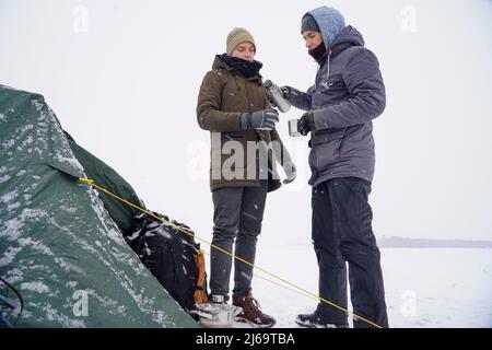 Les gars boivent le thé chaud des mugs thermo et versent l'eau d'un thermos. Ils se tiennent près de la tente pendant l'expédition d'hiver. Banque D'Images