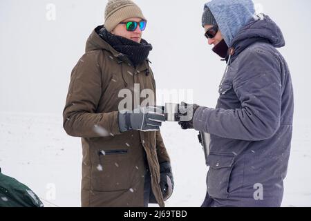 Les gars boivent le thé chaud des mugs thermo et versent l'eau d'un thermos. Ils se tiennent près de la tente pendant l'expédition d'hiver. Banque D'Images