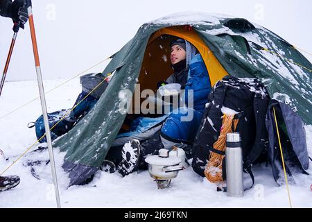 Un touriste s'assoit près d'une tente posée sur la neige, il sort un cordon d'un sac à dos pour une fixation supplémentaire de la tente pendant une tempête d'hiver Banque D'Images