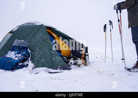 Un touriste s'assoit près d'une tente posée sur la neige, il sort un cordon d'un sac à dos pour une fixation supplémentaire de la tente pendant une tempête d'hiver Banque D'Images