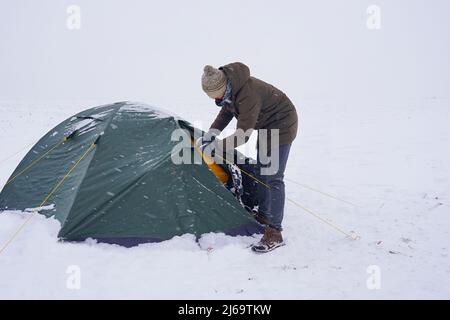 Un touriste s'assoit près d'une tente posée sur la neige, il sort un cordon d'un sac à dos pour une fixation supplémentaire de la tente pendant une tempête d'hiver Banque D'Images