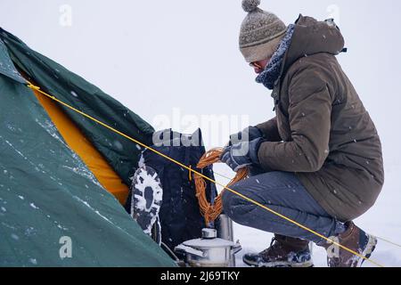 Un touriste s'assoit près d'une tente posée sur la neige, il sort un cordon d'un sac à dos pour une fixation supplémentaire de la tente pendant une tempête d'hiver Banque D'Images