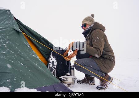 Un touriste s'assoit près d'une tente posée sur la neige, il sort un cordon d'un sac à dos pour une fixation supplémentaire de la tente pendant une tempête d'hiver Banque D'Images