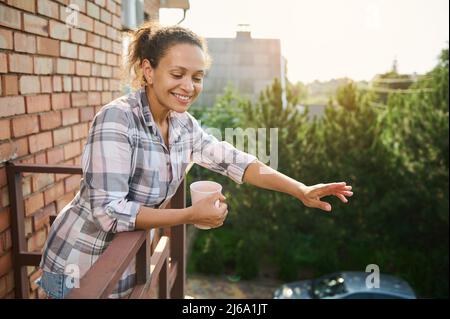 Une femme hispanique d'âge moyen souriante agite les mains saluant tout en se tenant sur le balcon d'une maison de campagne pendant une pause-café Banque D'Images