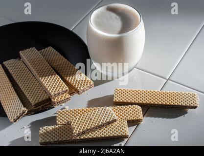biscuits de cachets sur une assiette noire et sur une table blanche, avec un verre de lait froid sur le côté éclairé par la lumière du soleil provenant d'une fenêtre Banque D'Images