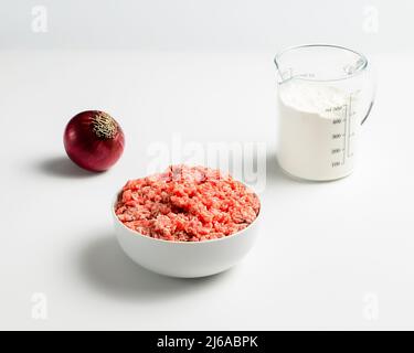 Assiette avec bœuf haché frais, oignon rouge, tasse graduée avec farine sur fond blanc, photo de stock Banque D'Images