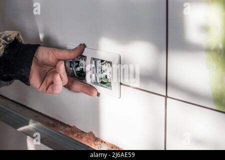 Un électricien installe un cadre en plastique sur une double prise sur un mur carrelé. Câblage électrique dans la maison, réparation. Banque D'Images