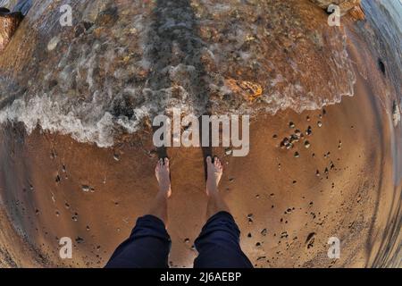 Première personne perspective Fisheye des pieds de l'homme dans l'eau peu profonde avec des vagues qui se roulent dans Sandy Beach Banque D'Images