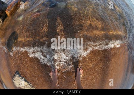 Première personne perspective Fisheye des pieds de l'homme dans l'eau peu profonde avec des vagues qui se roulent dans Sandy Beach Banque D'Images