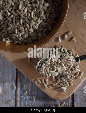 Graines de tournesol séchées et salées sur une cuillère et dans un bol sur une planche à découper en bois sur une table rustique éclairée par la lumière du soleil venant d'une fenêtre. Banque D'Images