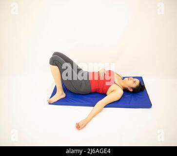 belle femme orientale blanc athlétique reposant dans des vêtements de sport gris et un chemisier rouge, avec ses jambes rétractées sur un tapis bleu, sur fond blanc Banque D'Images