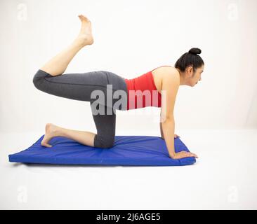 belle femme blanche orientale en vêtements de sport gris et blouse rouge, s'étirant sur la jambe droite, sur un tapis bleu, sur un fond blanc Banque D'Images