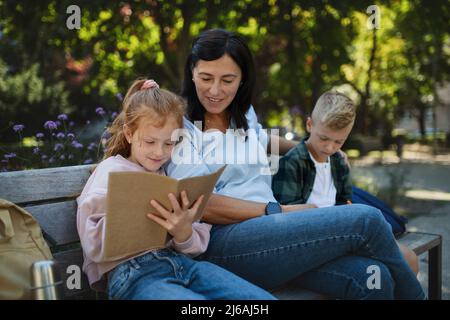 Une femme âgée heureuse avec des petits-enfants assis à son banc et aidant à faire ses devoirs à l'extérieur dans le parc. Banque D'Images