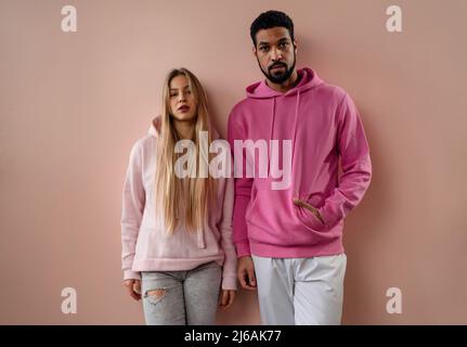 Portrait de studio de mode de jeune couple biracial dans des sweats à capuche posant sur fond rose. Banque D'Images