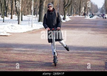 Le gars fait un scooter électrique au printemps dans le parc. Il porte une veste et un chapeau chauds. À proximité, il fait neige et froid Banque D'Images