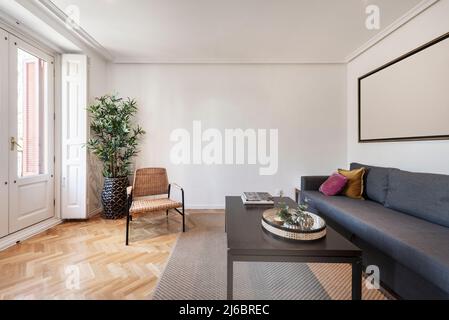 Séjour avec canapé en tissu, table en bois sombre, parquet en chêne à chevrons, plantes décoratives, chaise en osier et balcon avec volets en bois blanc Banque D'Images