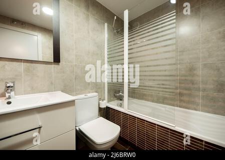 Salle de bains avec lavabo en résine blanche sur armoire en bois avec tiroirs, toilettes blanches, miroir sur le mur, cabine de douche en verre et carreaux bruns Banque D'Images