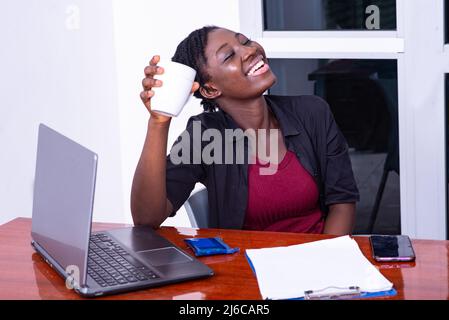 une jeune femme d'affaires riante assise au bureau avec un ordinateur tenant une tasse de café Banque D'Images
