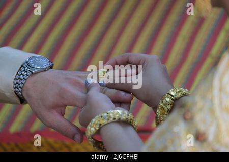 Mariage thaïlandais : une mariée somptueusement décorée est en train de mettre un anneau doré au doigt du marié Banque D'Images