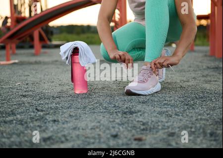 Gros plan d'une athlète féminine, sportswoman nouant des lacets avant un entraînement intensif sur des appareils de gym sur le terrain de sport d'un parc de la ville pendant un été chaud d Banque D'Images