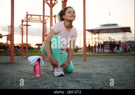 Belle sportive afro-américaine d'âge moyen, motivée, nouant des lacets de chaussures, sur un terrain de sport en plein air contre des machines de gym en plein air au coucher du soleil Banque D'Images