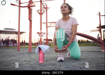 Sportif multiethnique d'âge moyen déterminé qui noue des lacets, debout sur un terrain de sport extérieur contre une barre transversale et des appareils de gym au coucher du soleil Banque D'Images