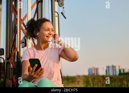 Femme afro-américaine d'âge moyen gaie dans des vêtements de sport, portant des écouteurs et écoutant de la musique, sourit en regardant de côté en se reposant entre les ensembles d Banque D'Images