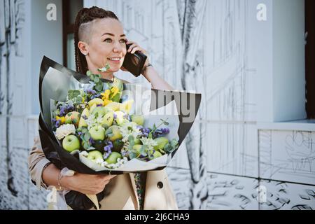 Livraison de fleurs. Jeune femme moderne taille basse avec téléphone portable recevant de belles fleurs bouquet de livraison à l'extérieur Banque D'Images
